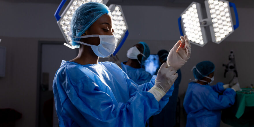 African american female doctor with face mask wearing protective gloves in hospital operating room. Medicine, healthcare and medical services, unaltered.